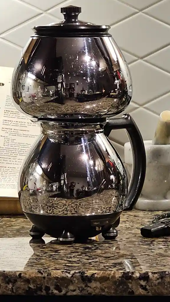A glass coffee maker sits on a granite countertop, its reflective surface mirroring the kitchen and a person taking the…