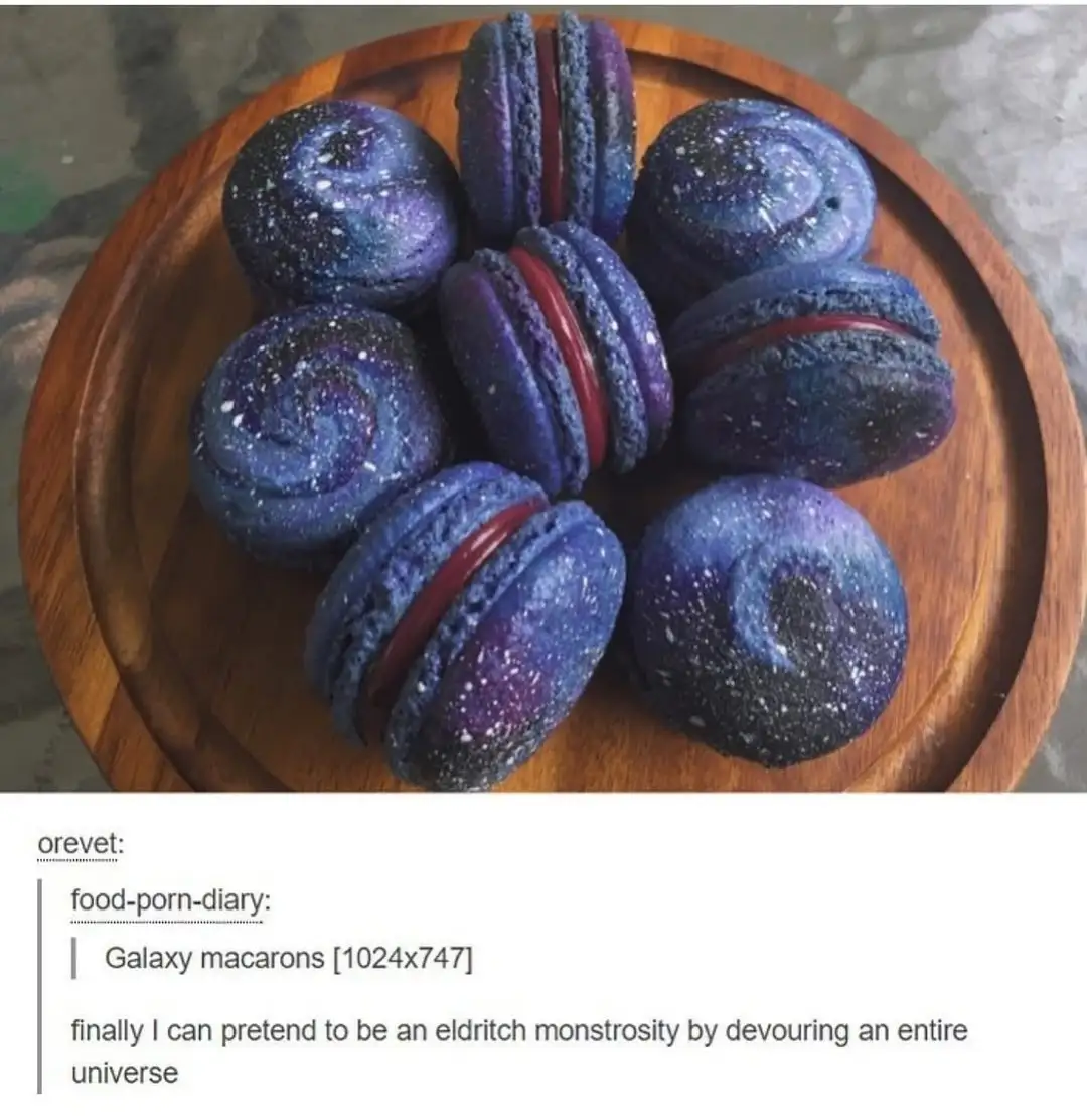 A wooden plate holds several macarons decorated to resemble galaxies, with swirling blue, purple, and black colors and white…