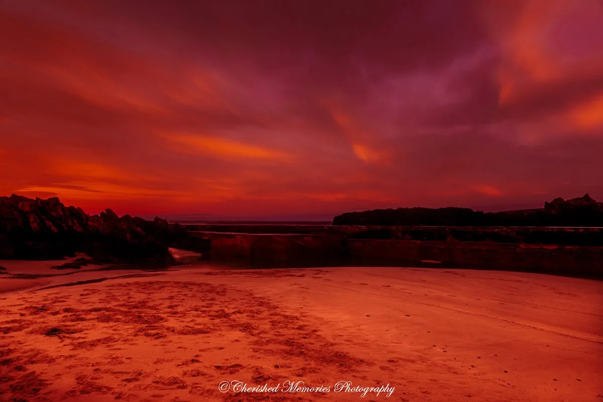 A dramatic, long-exposure photograph of a beach at sunset, where the sky and sand are bathed in intense red and orange hues…