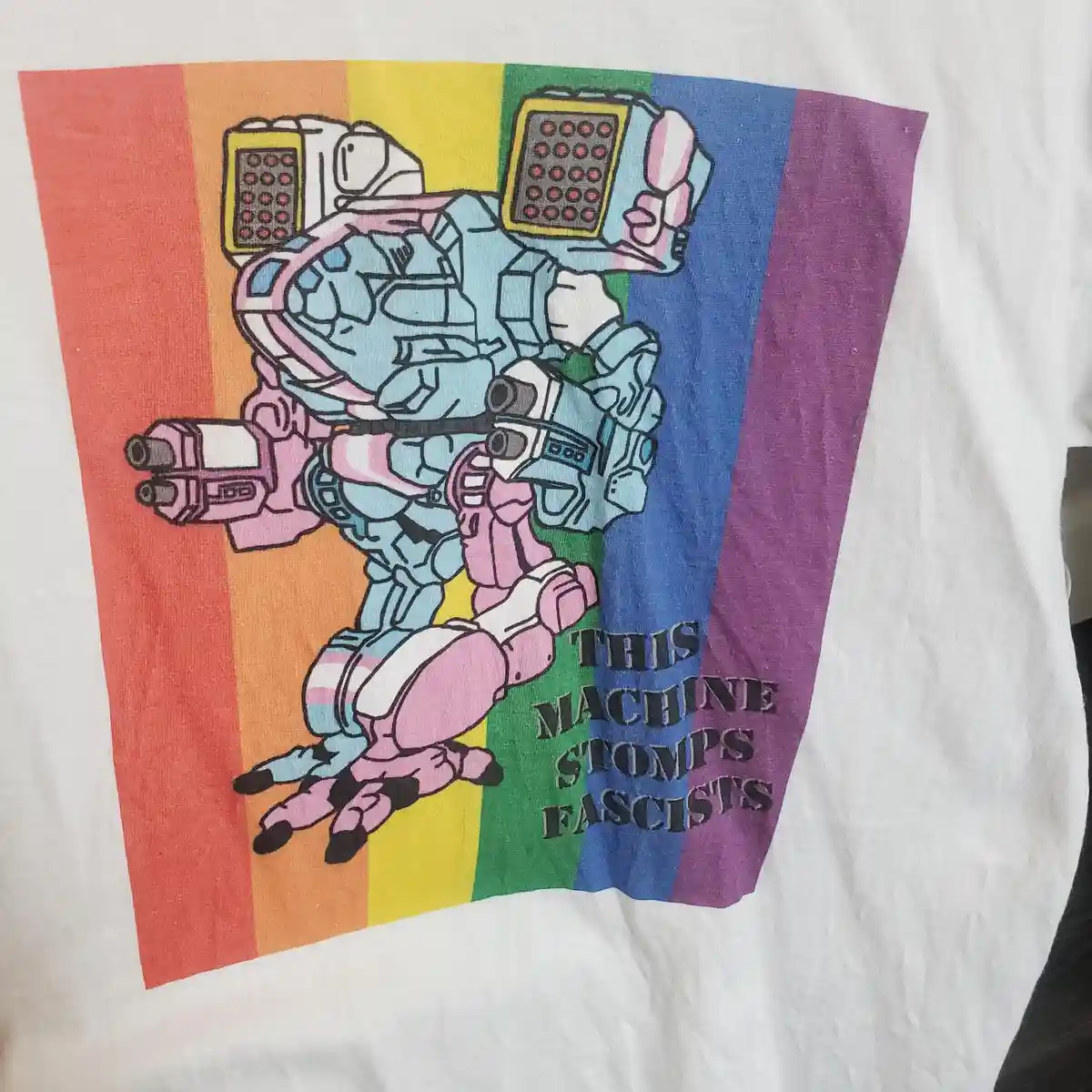 A white t-shirt features a cartoon robot with blue and pink armor, holding weapons, set against a rainbow background. The…