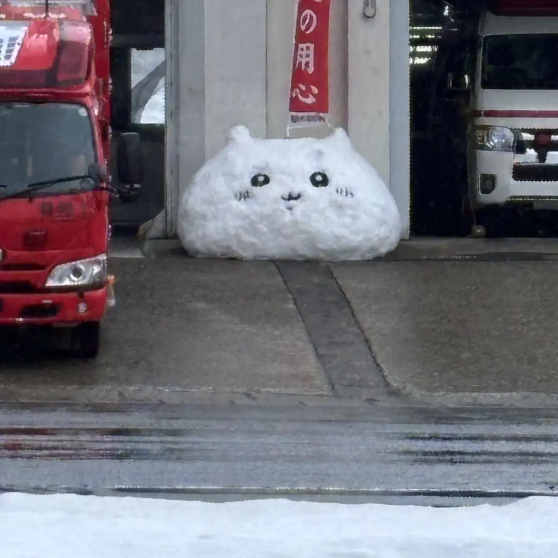 A large, fluffy snowball shaped like a cute cat with drawn-on black eyes, nose, and whiskers sits on a sidewalk between a…