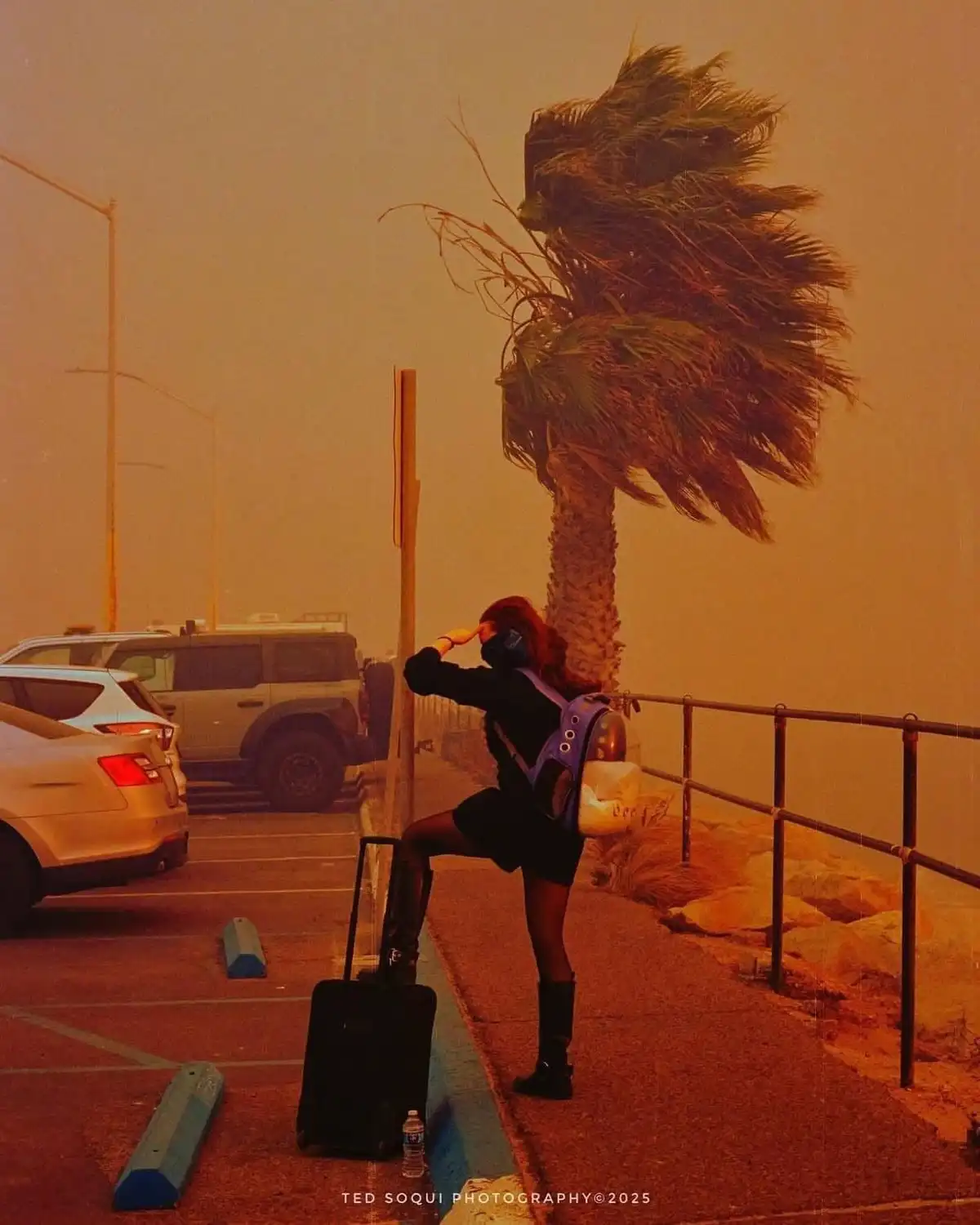 A person wearing a face mask and carrying a backpack stands in a parking lot next to a suitcase, saluting with one hand…