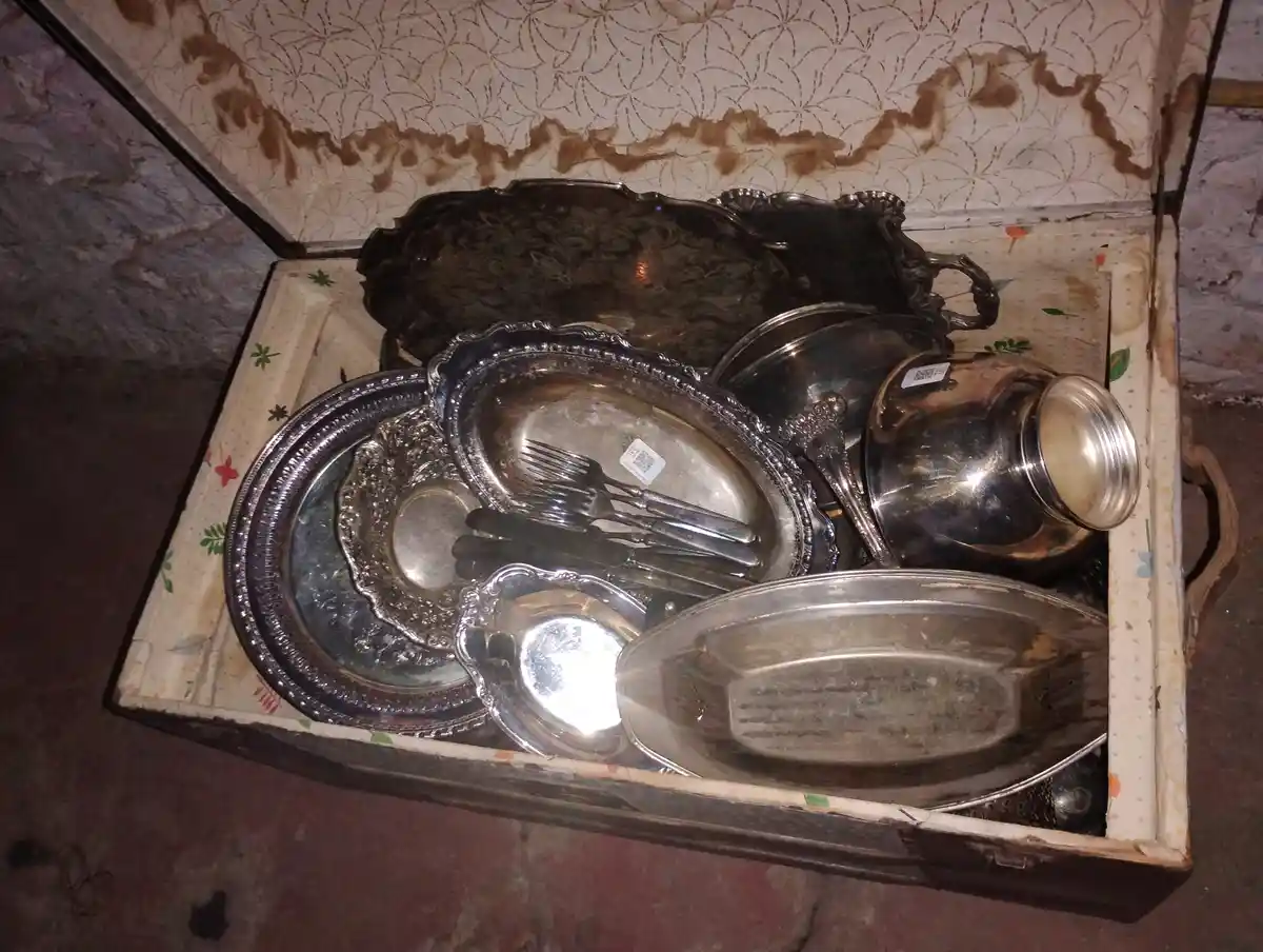 An open, worn wooden chest contains a jumble of tarnished silver platters, bowls, and cutlery, all piled together with…