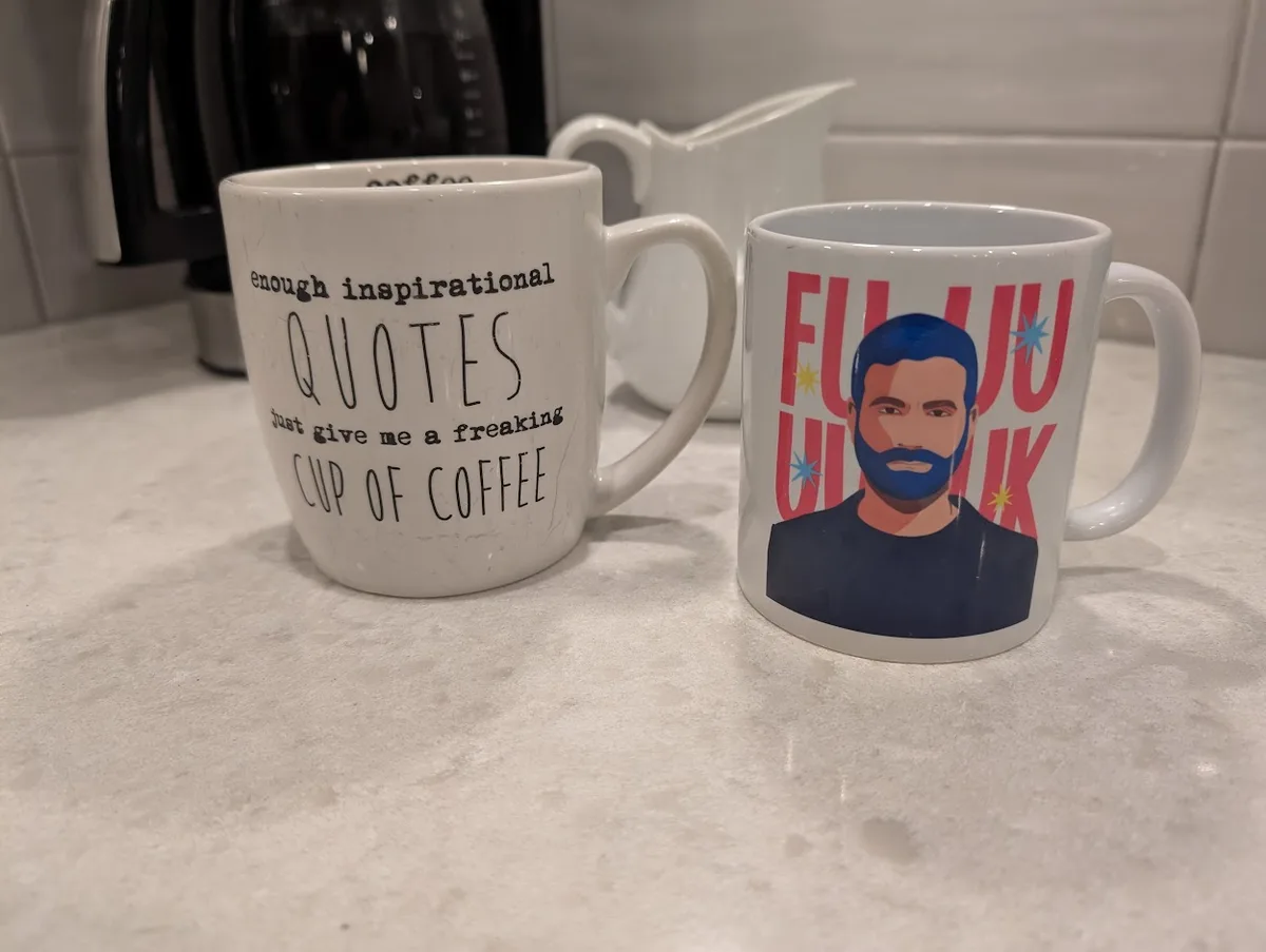 Two coffee mugs sit on a kitchen counter. The left mug has black text reading 'enough inspirational QUOTES just give me a…
