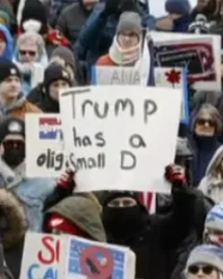 A person in a black hooded jacket and red gloves holds a white sign that reads 'Trump has a small D' at a protest. Other…