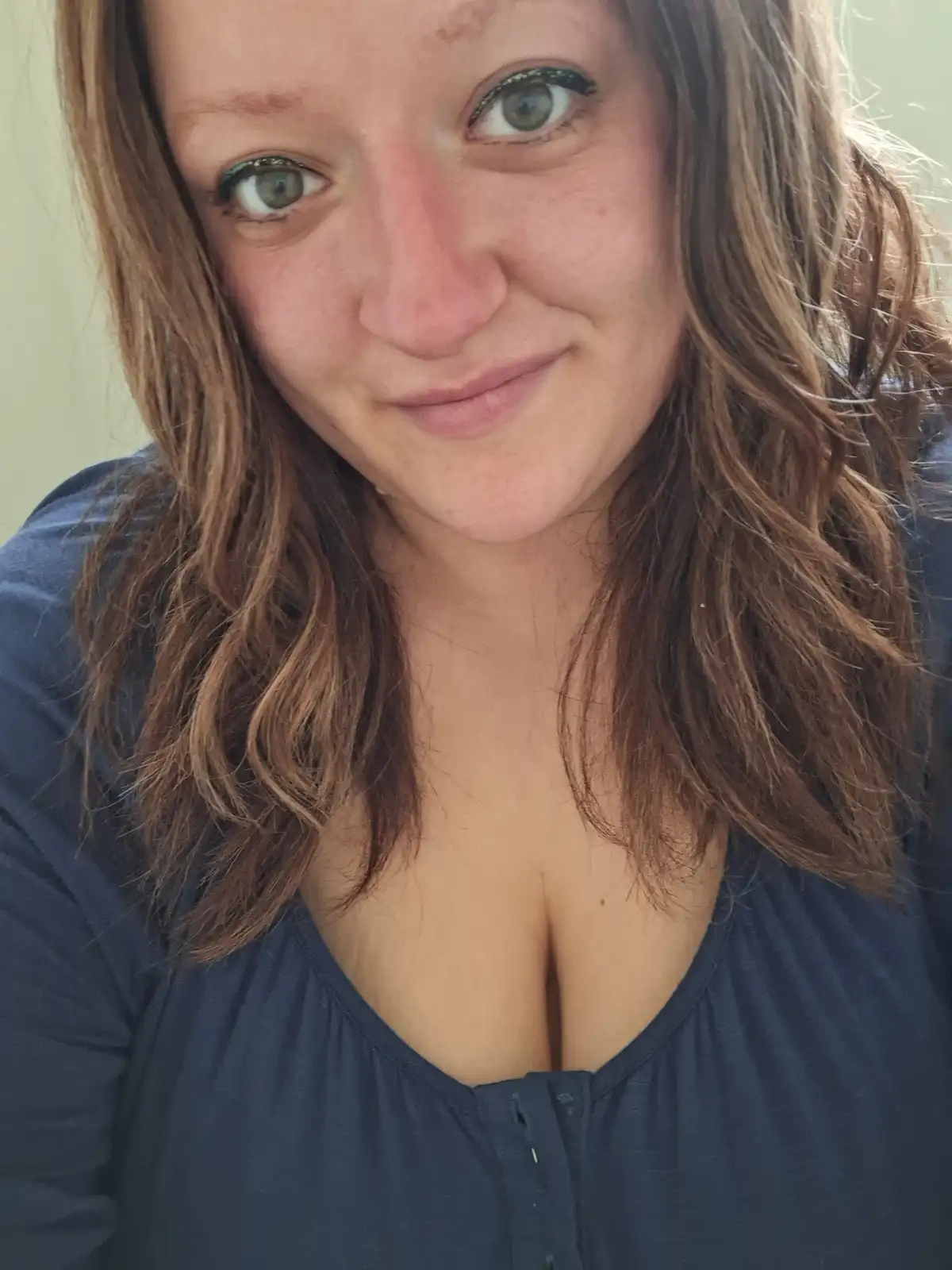 A close-up selfie of a woman with brown hair and green eyes, wearing a dark blue top with a low neckline. She has a slight…
