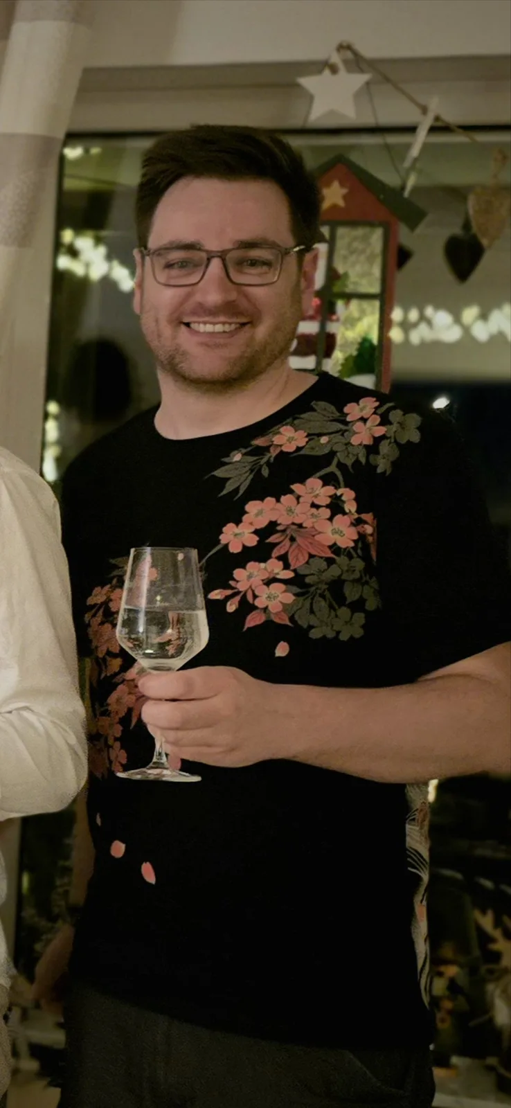 A man with glasses and a beard smiles while holding a wine glass filled with clear liquid, wearing a black t-shirt with a…