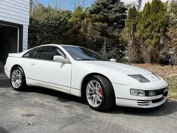 a white nissan 300zx sports car is parked on an asphalt driveway in front of a house with a garage door and various green…