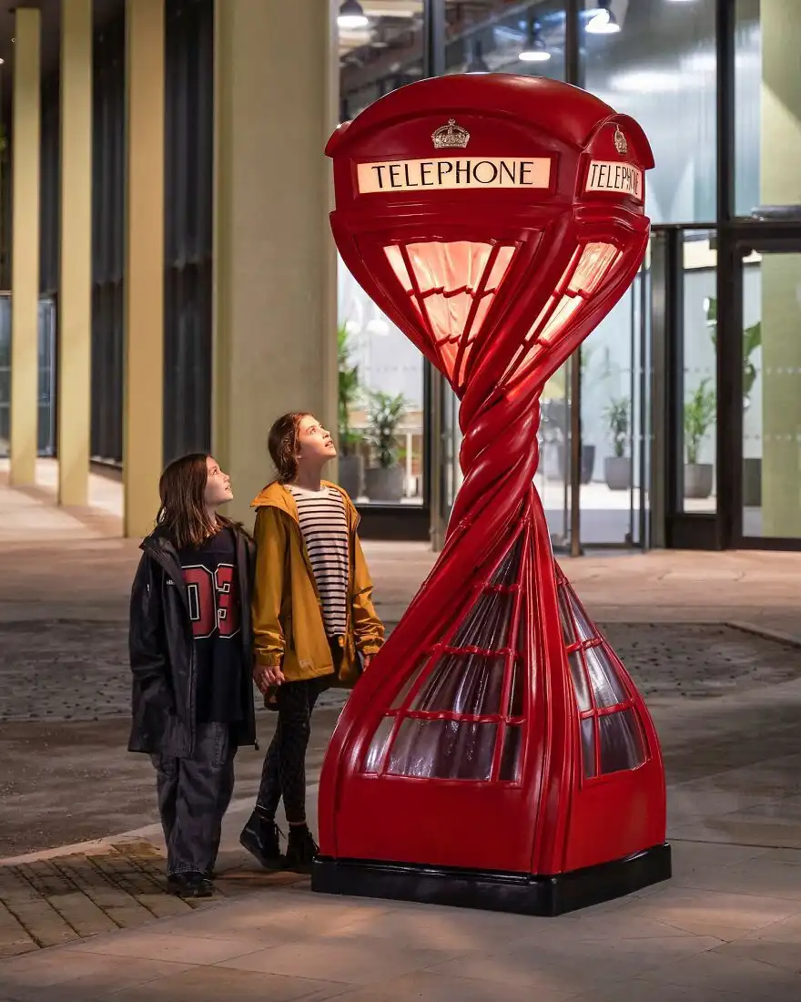 Two children stand looking up in awe at a giant, twisted red telephone booth sculpture that spirals downward, with the word…