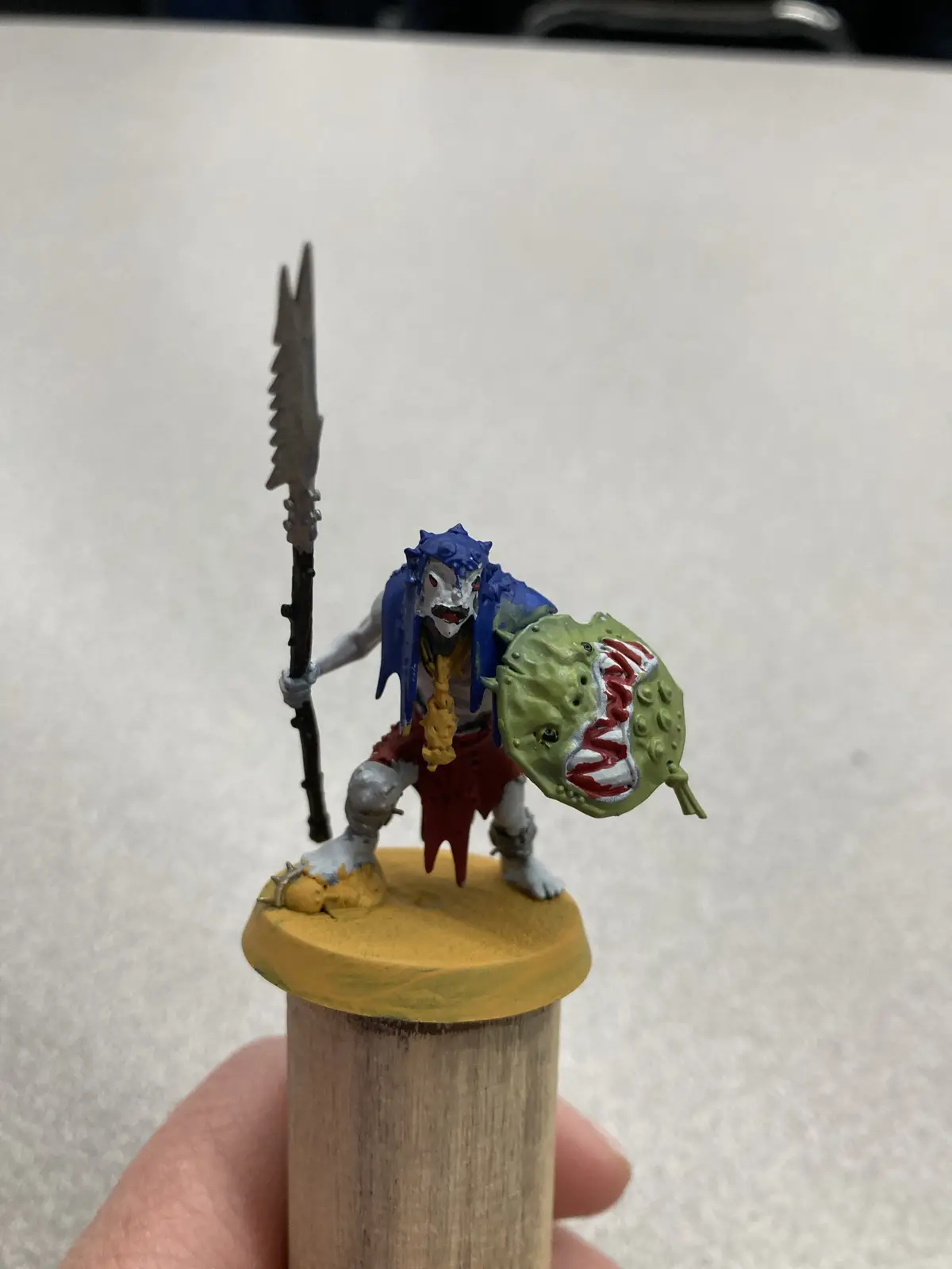 A hand holds a painted miniature warrior figurine on a wooden dowel, featuring a blue-haired, white-faced character with red…