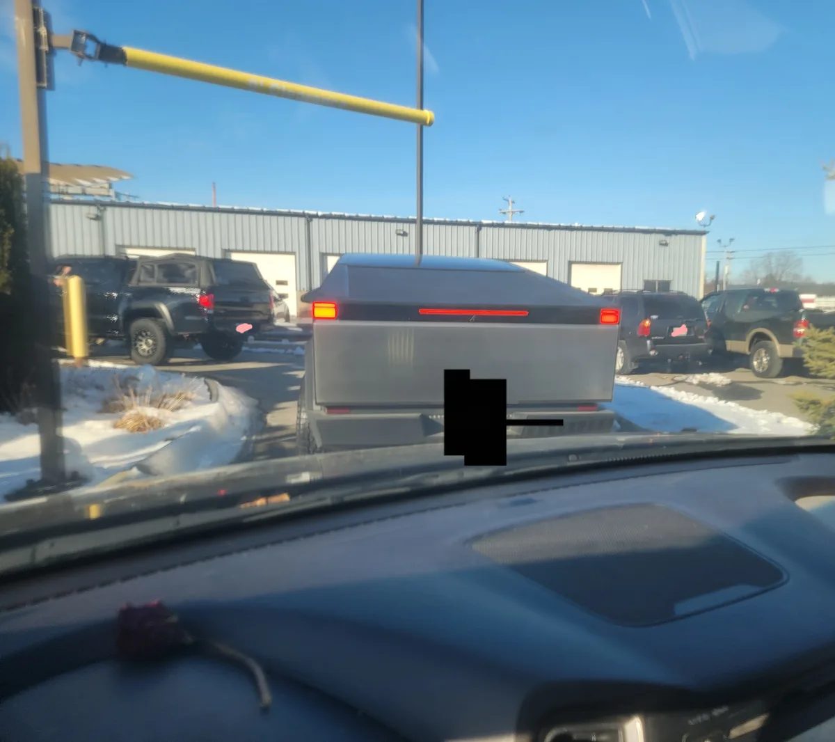 A photo taken from inside a car shows a silver Tesla Cybertruck stopped at a drive-thru with a yellow clearance bar…
