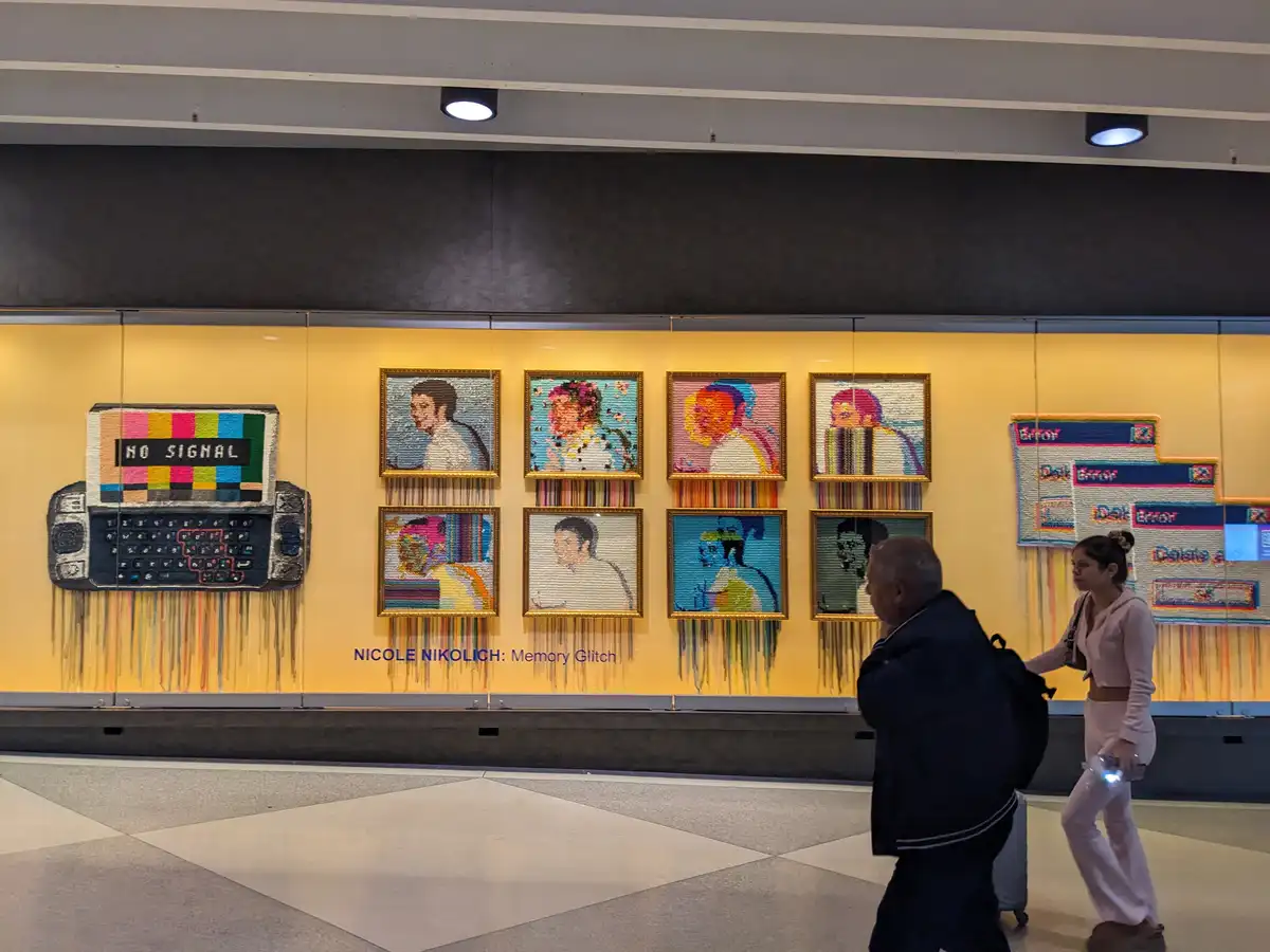 A photo of a public art installation titled 'Memory Glitch' by Nicole Nikolich, featuring colorful pixelated portraits and a…