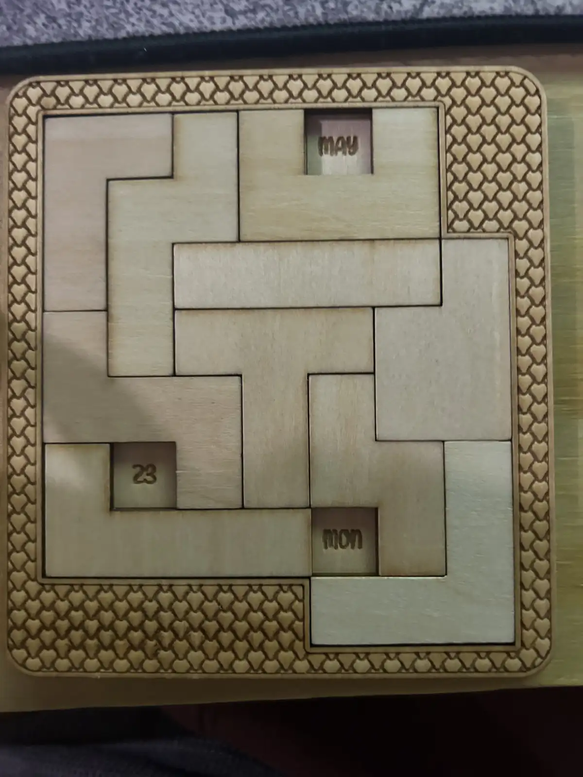 A wooden puzzle calendar with interlocking pieces, showing the date 'may 23' and the day 'mon' in engraved slots, arranged…