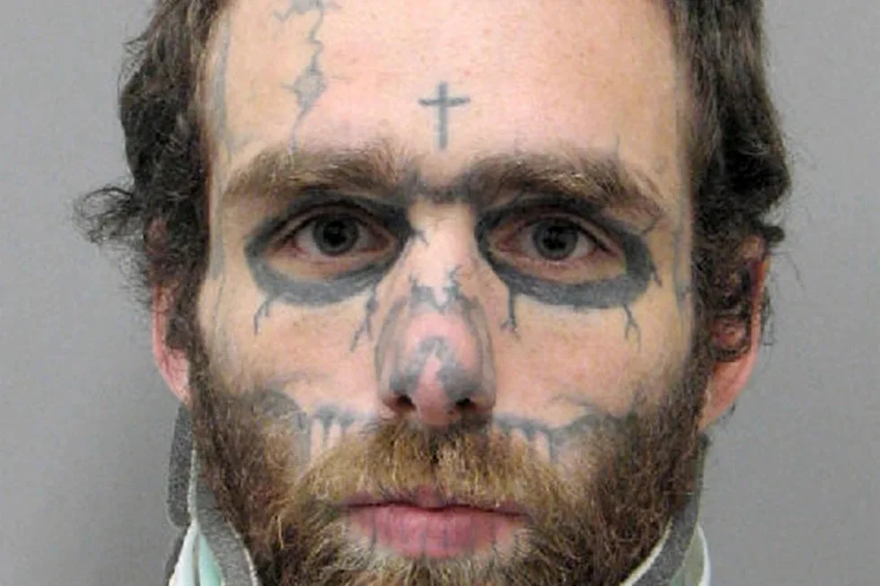A mugshot-style photo of a man with dark, crude tattoos drawn on his face resembling a skull and crossbones, including a…