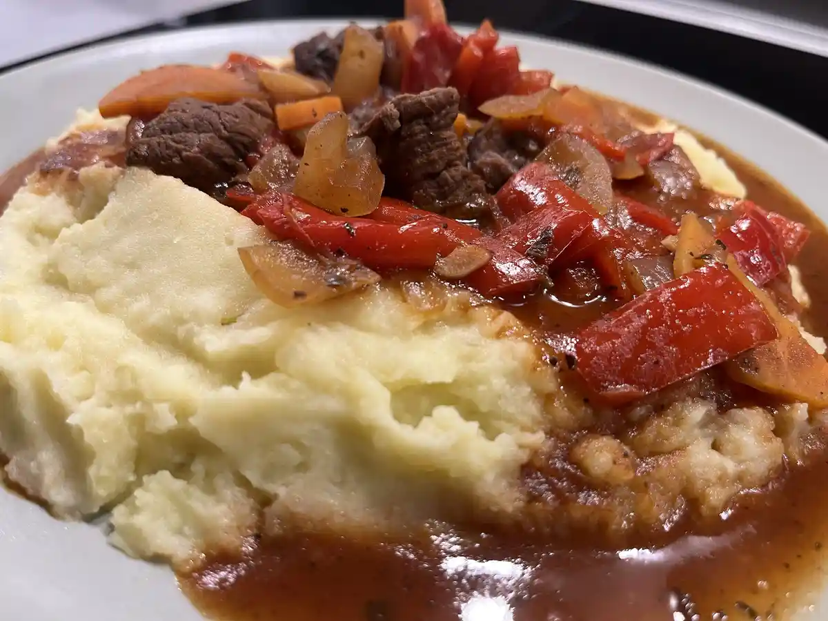 A close-up photo of a white plate holding a serving of mashed potatoes topped with a stew containing chunks of beef, red…