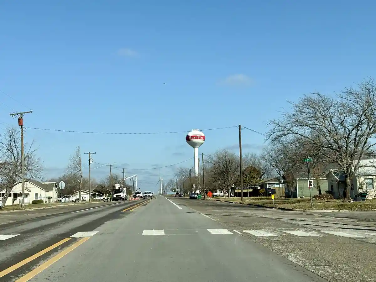 A photo of a quiet rural road with houses on both sides, bare trees, and a white water tower in the distance with the name…