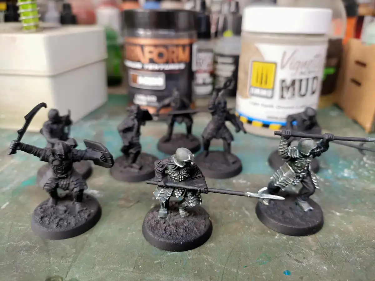 Several unpainted or partially painted fantasy miniatures, mostly in black, stand on circular bases on a green work surface…