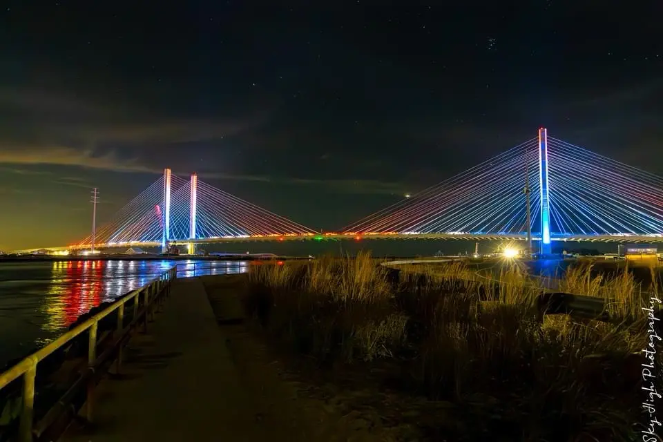 A nighttime photograph of a modern cable-stayed bridge illuminated with colorful lights against a starry sky, viewed from a…