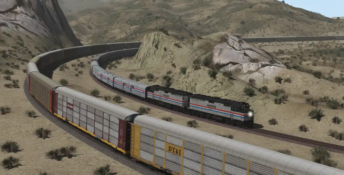 A screenshot from a train simulation game shows two long trains curving around a desert canyon. One train, led by Amtrak…
