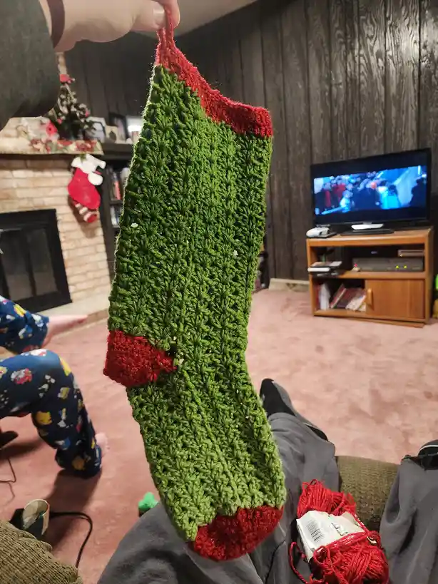 A person holds up a crocheted green stocking with red trim, which has a noticeable hole near the middle. The background…