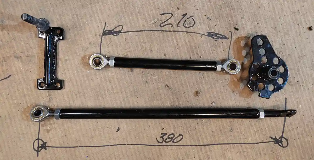 Two black metal suspension control arms lie on a cardboard surface, one marked with a hand-drawn measurement of 210 and the…