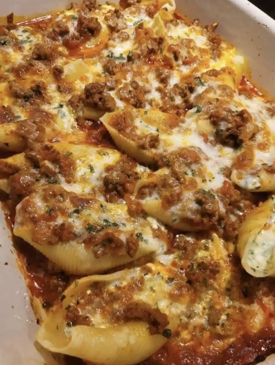 A close-up photo of a casserole dish filled with baked pasta shells covered in melted cheese, meat sauce, and herbs. The…