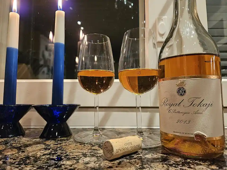 A bottle of Royal Tokaji 6 Puttonyos Aszu 2013 wine sits next to two filled glasses on a granite countertop, with a cork…