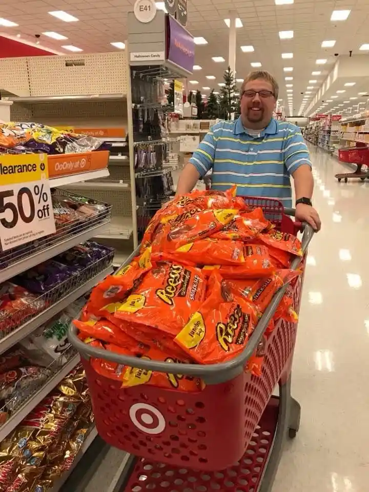 A man with glasses and a striped polo shirt pushes a red Target shopping cart overflowing with orange Reese's Peanut Butter…