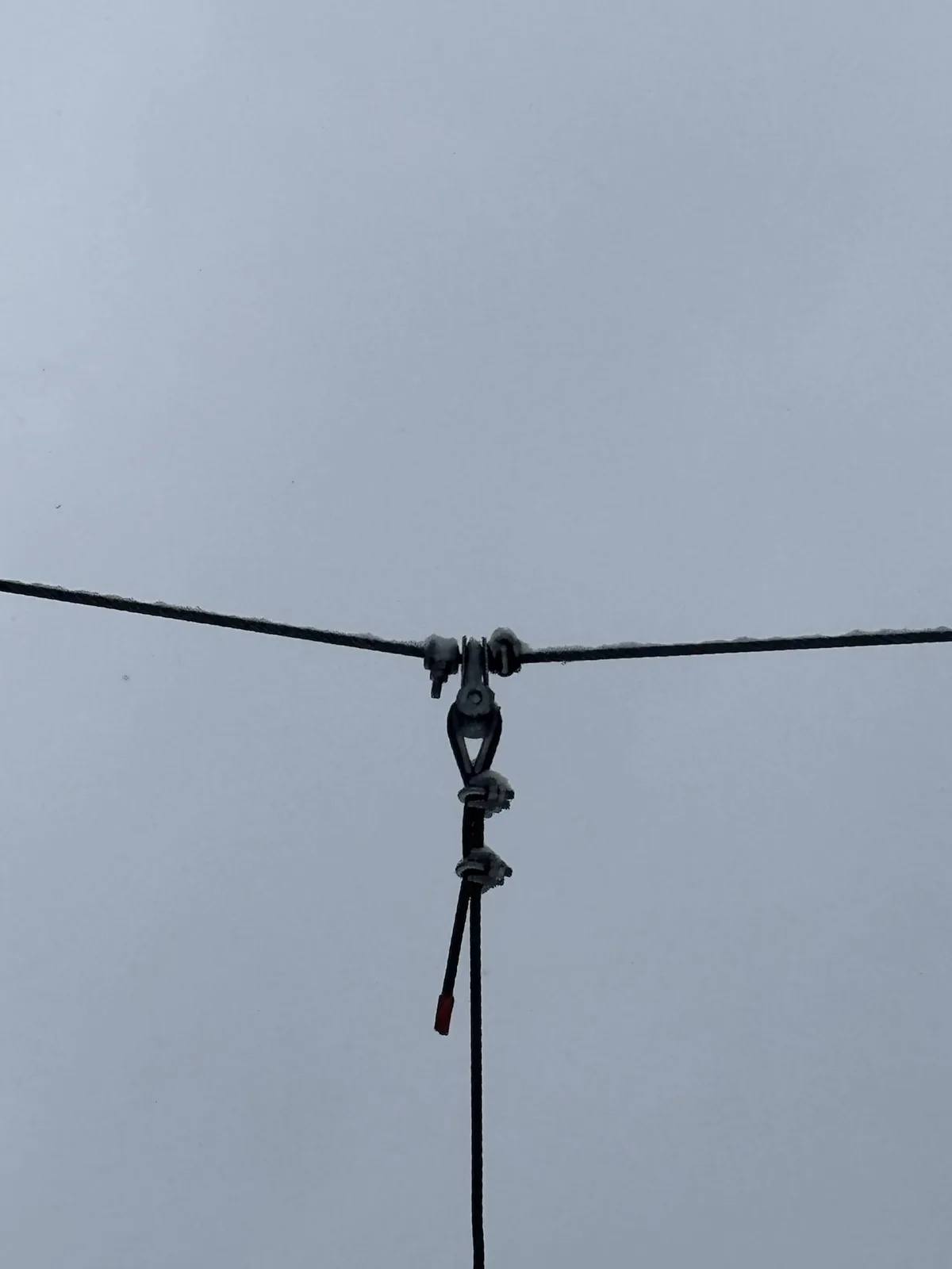 A close-up photo shows a metal ski lift cable pulley mechanism covered in snow against a gray sky. A vertical cable hangs…