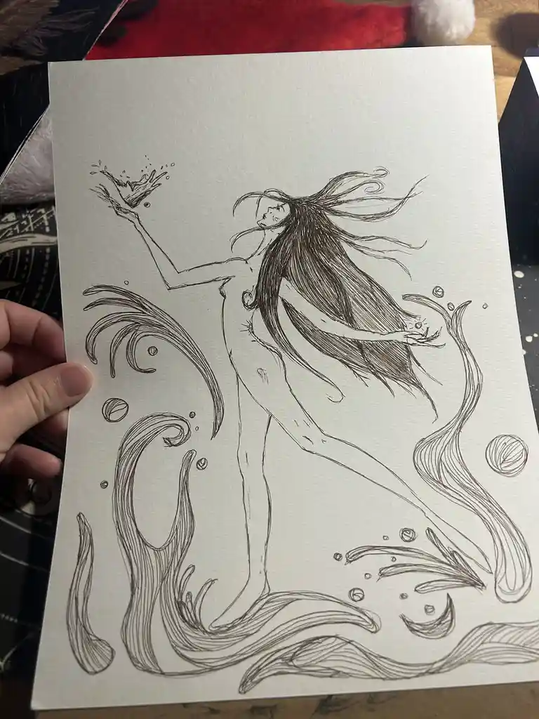 A hand holds up a black-and-white ink drawing of a mermaid with long flowing hair, her upper body human and lower body…
