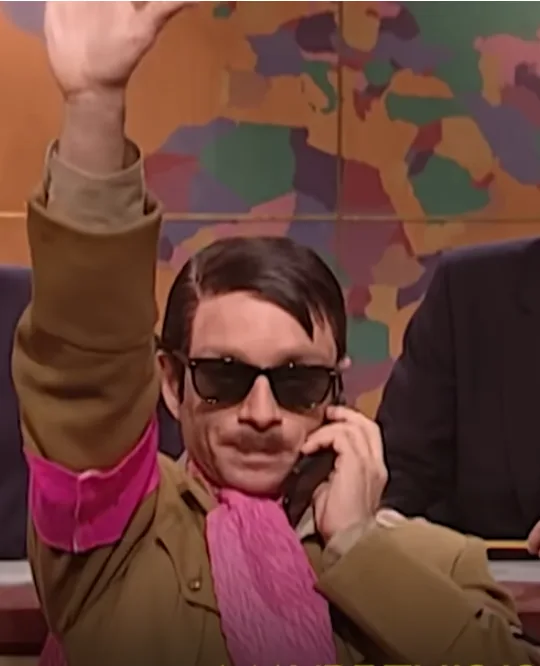 a man wearing sunglasses, a brown jacket with a bright pink shirt and scarf, holds a phone to his ear while raising his…