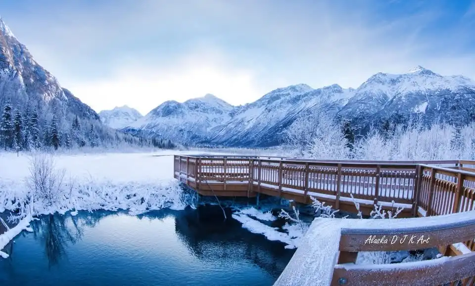 A serene winter landscape featuring a wooden boardwalk extending over a partially frozen lake, surrounded by snow-covered…