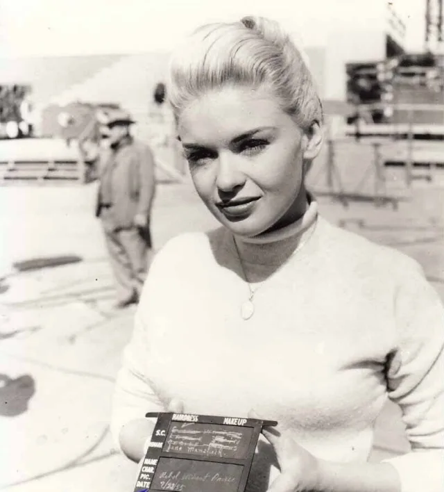 A black-and-white photo of a blonde woman in a turtleneck holding a film slate that reads 'S.C. SHOT WITHOUT CARRIE'. The…