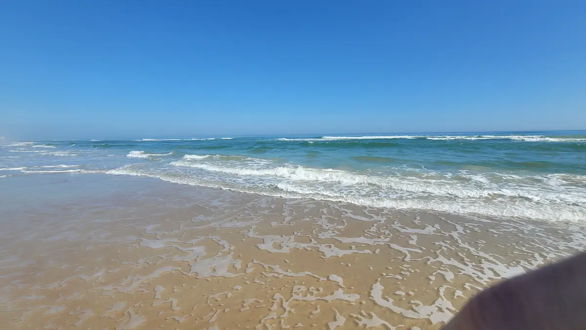 A serene beach scene with turquoise ocean waves rolling onto wet, sandy shores under a clear blue sky. The image has text…