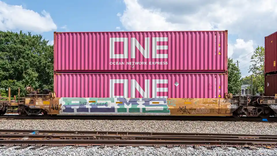 Two stacked pink shipping containers branded with 'ONE OCEAN NETWORK EXPRESS' sit atop a rusty train car covered in…