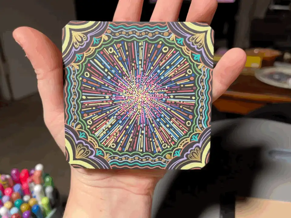 A hand holds a square coaster with a colorful, intricate mandala-like design featuring radial patterns and multiple hues…