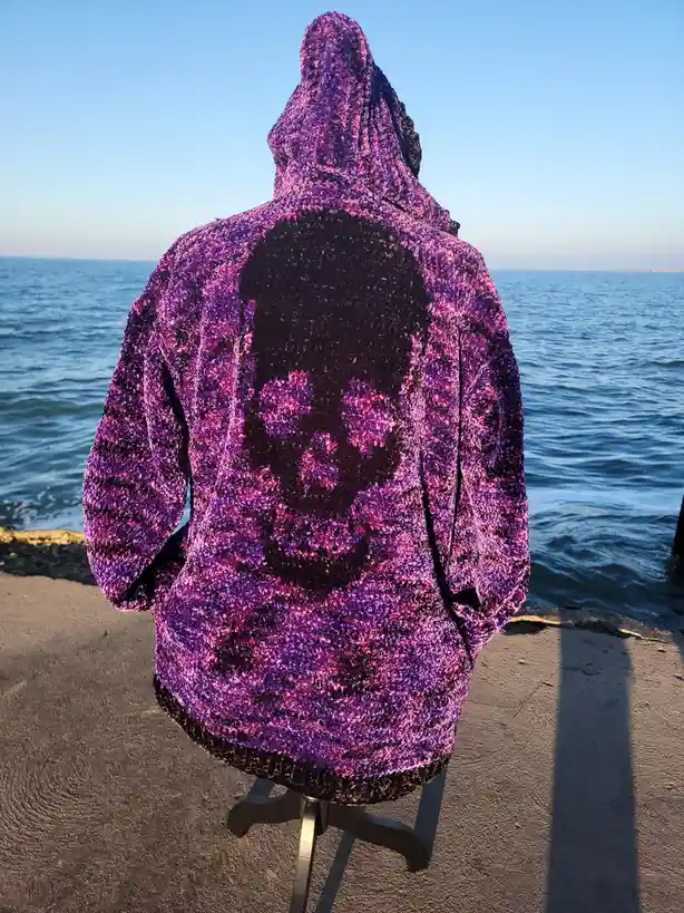 A person wearing a purple and pink textured hooded jacket with a large black skull design on the back, standing with their…
