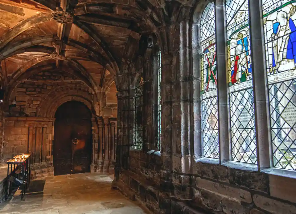 An image of a dimly lit, stone-walled church corridor with arched ceilings, stained glass windows depicting saints, and a…