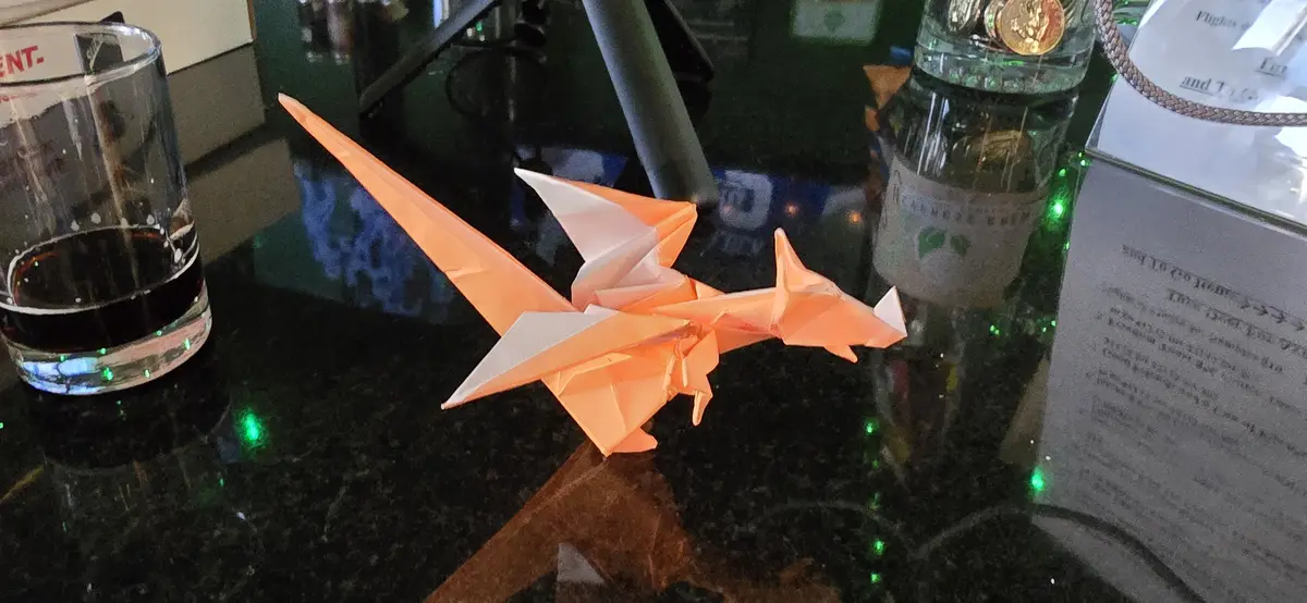 An orange and white origami dragon sits on a dark, reflective countertop next to a glass of dark liquid, a bottle, and a…