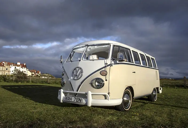 A vintage cream and white Volkswagen Type 2 van is parked on a grassy field under a dramatic, cloudy sky. The vehicle…
