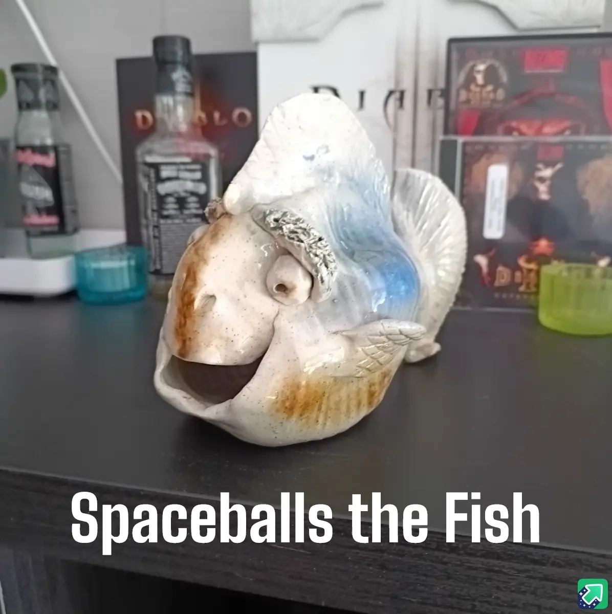 A ceramic fish figurine with a wide, open mouth and a comically large, spiky dorsal fin sits on a dark shelf. Behind it are…