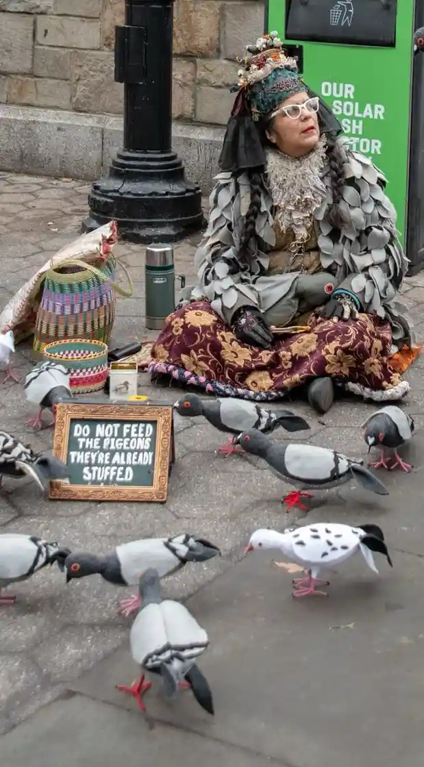 A person dressed in elaborate, layered clothing with a crown and glasses sits on a patterned blanket on a sidewalk…