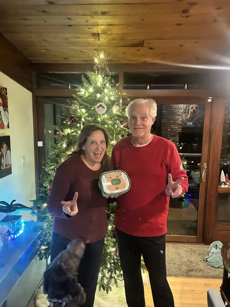 A man and woman stand in front of a decorated Christmas tree, both making peace signs with their hands. The woman holds a…