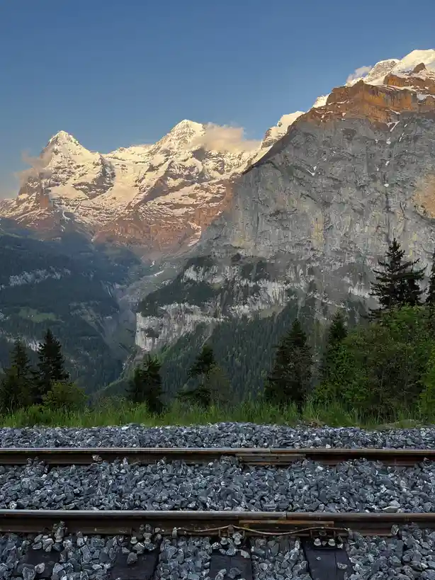A scenic photograph shows a railway track in the foreground, leading toward majestic snow-capped mountains under a clear…