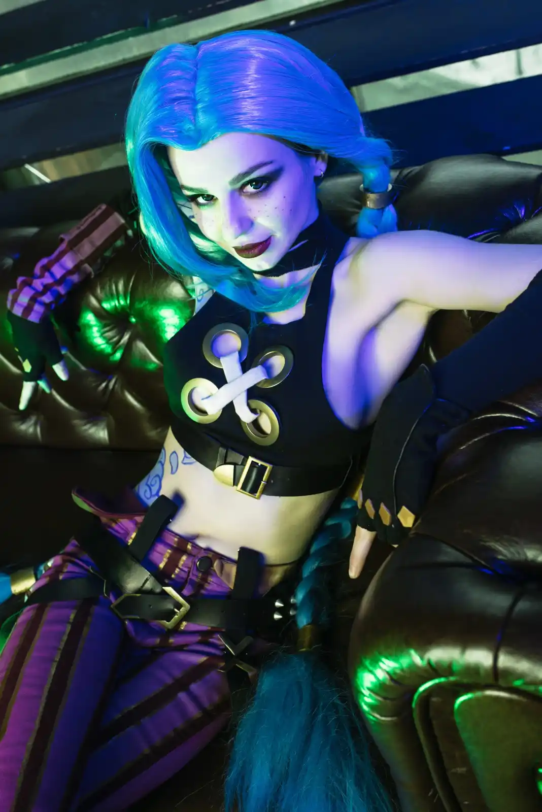 A person in a cosplay of the character Jinx from League of Legends sits on a brown leather couch, looking directly at the…