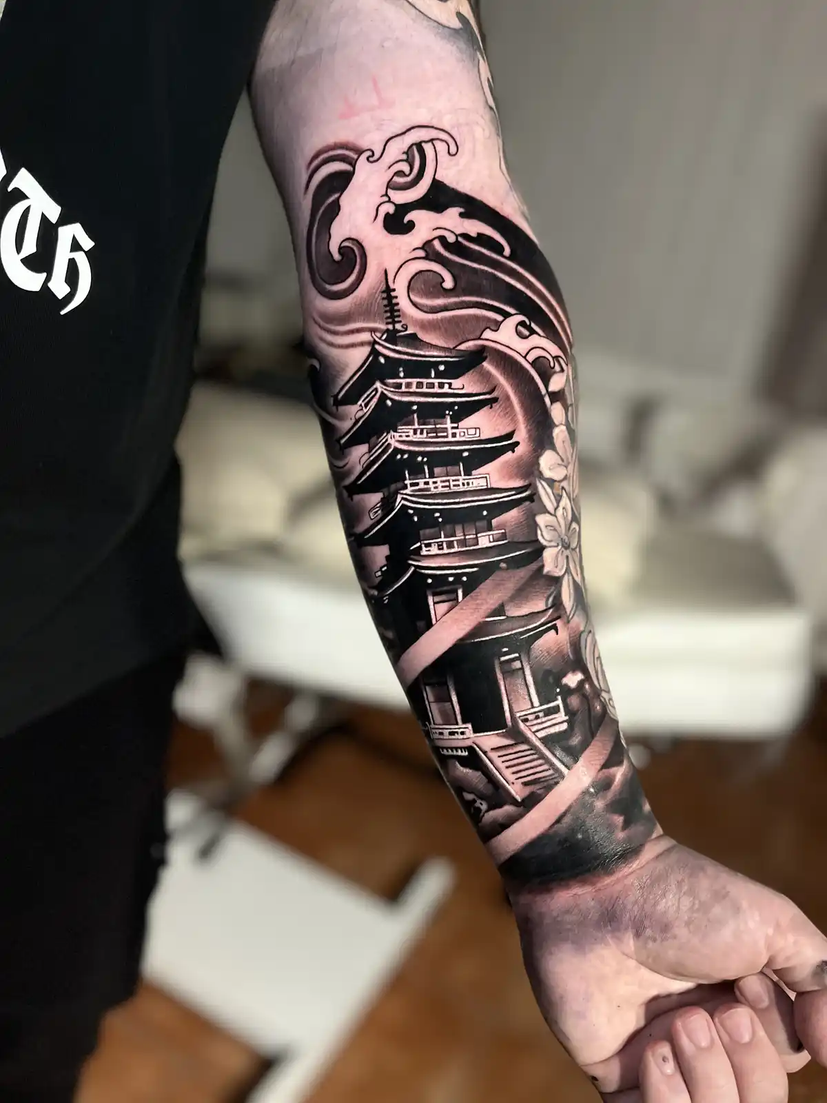A person's forearm displays a detailed black and grey tattoo of a traditional Japanese pagoda surrounded by stylized waves…