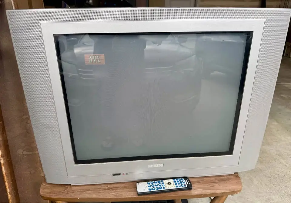 A silver Philips CRT television with a blank, slightly reflective screen showing a faint 'AV2' label, sitting on a wooden…