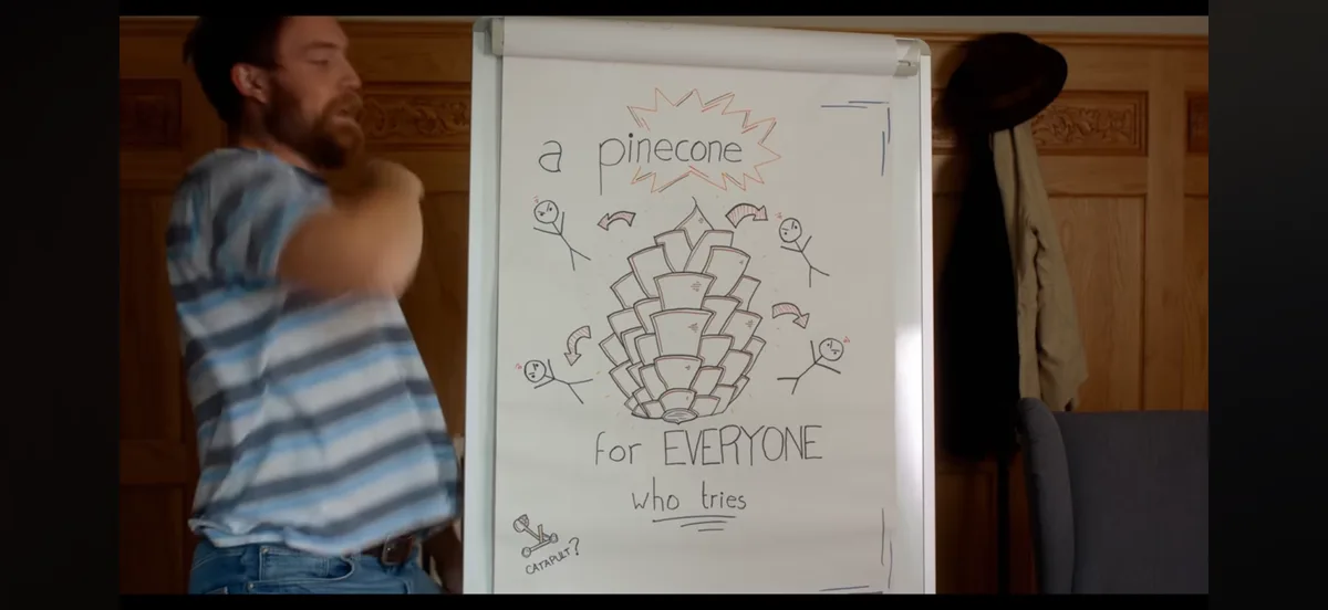 A man in a striped shirt stands beside a whiteboard with a hand-drawn diagram showing a large pinecone surrounded by stick…