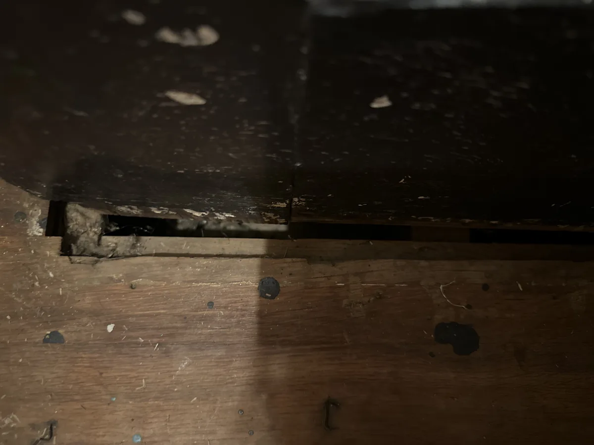 A close-up photo shows a dark, worn wooden floor with a visible gap between two floorboards, revealing a darker space…