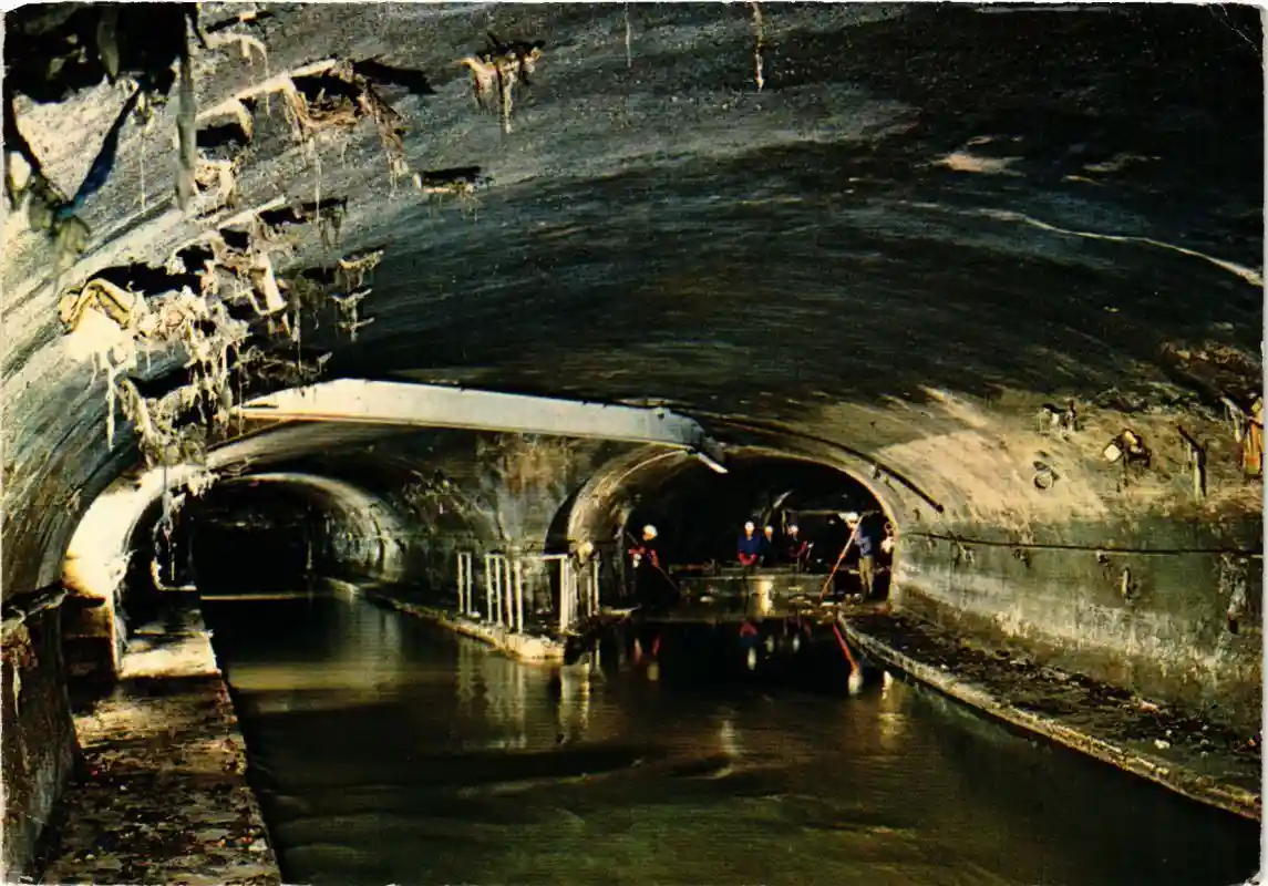 A dark, wet, arched tunnel with crumbling, dripping walls and a water-filled floor. Several workers in hard hats are visible…